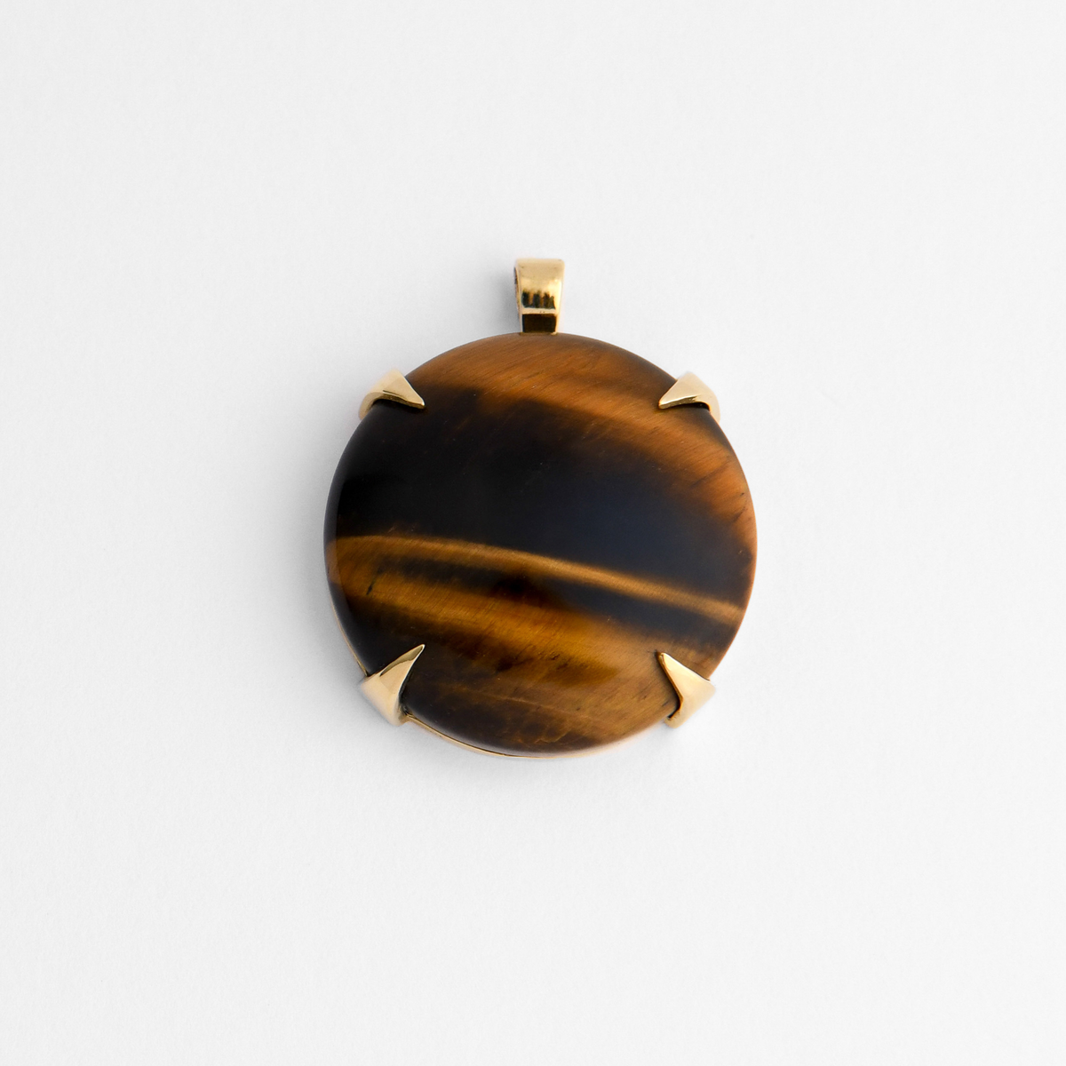 Spar, a round pendant in warm, golden-yellow brass, with four triangular claws holding golden-brown Tiger’s Eye, with dark, almost metallic bands that shift in the light. As a natural stone, each piece varies.