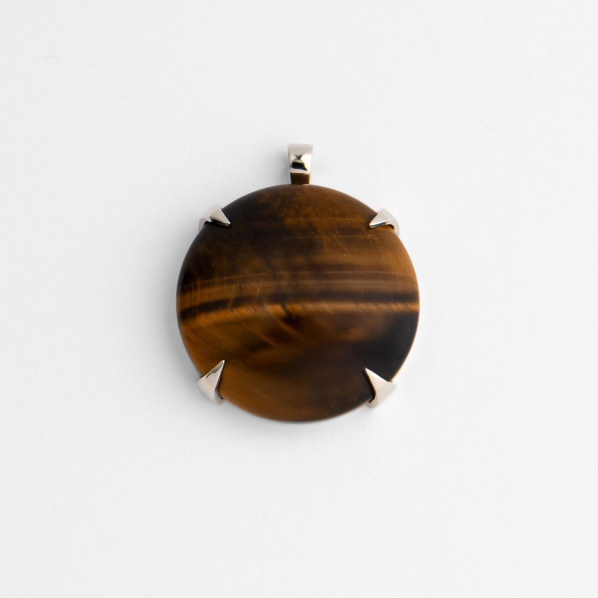 Spar, a round pendant in cool, silvery white bronze, with four triangular claws holding golden-brown Tiger’s Eye, with dark, almost metallic bands that shift in the light. As a natural stone, each piece varies.