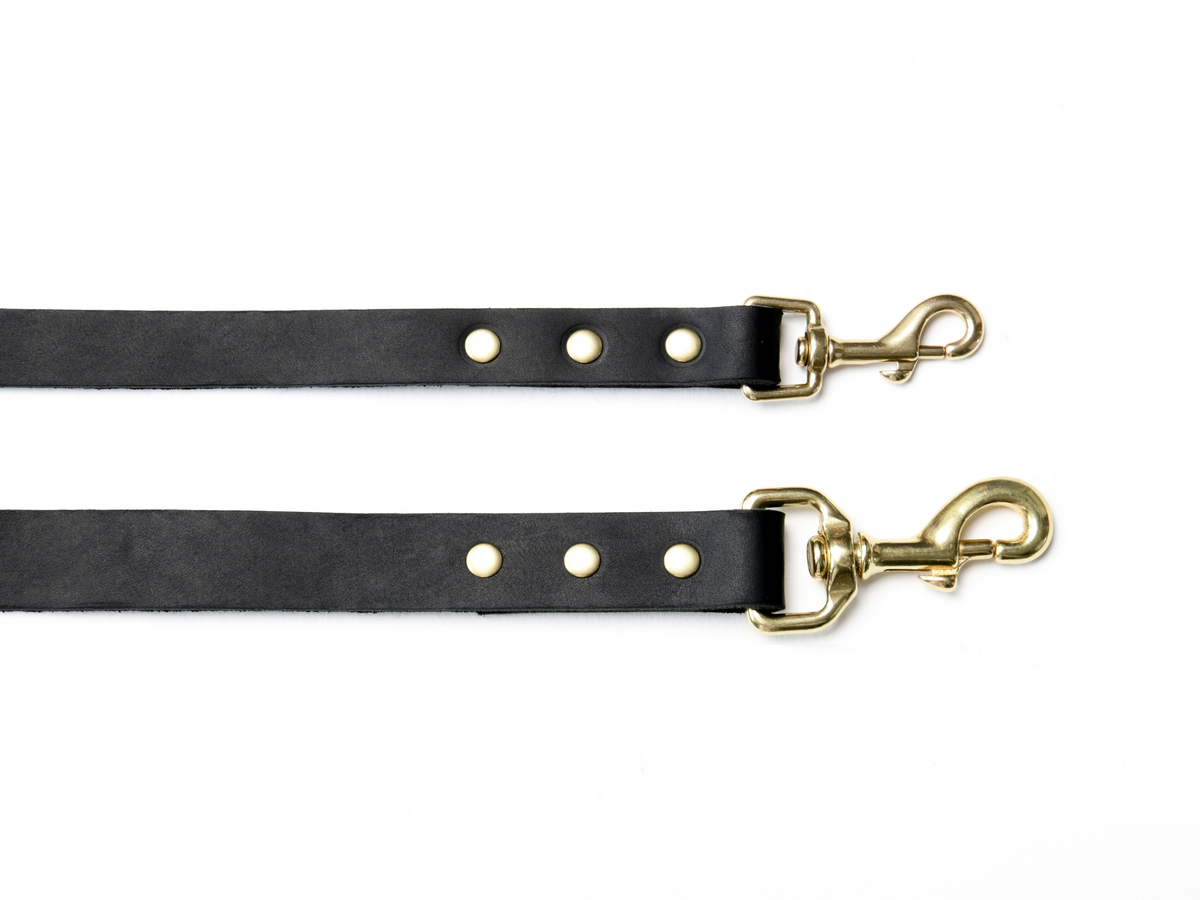 Two black cowhide leather dog leads in XS/S and M/L sizes, shown together to compare their widths and sizes of the brass trigger clips. Each clip is secured with three brass studs.