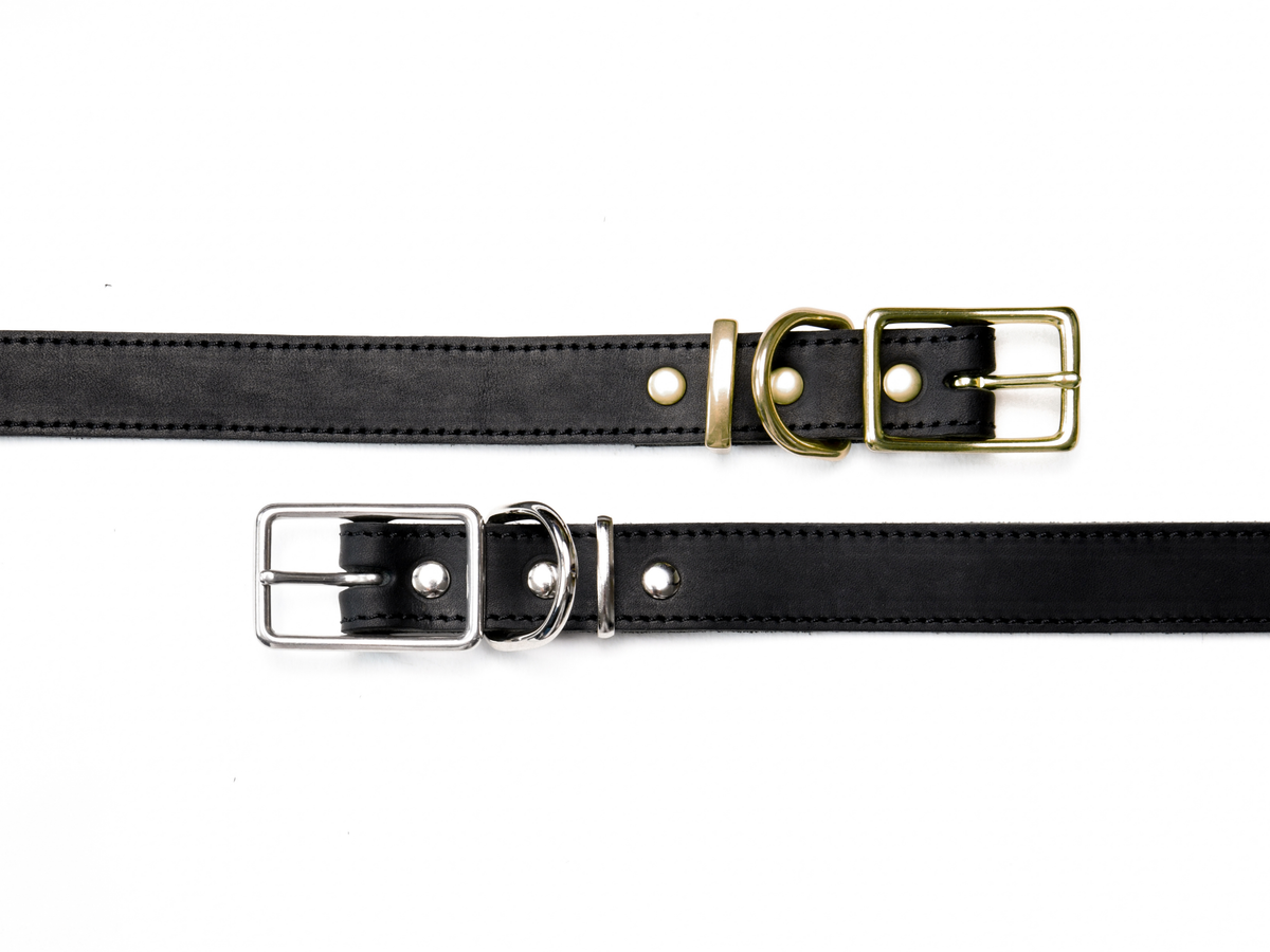 Two black cowhide leather dog collars laid flat to compare the hardware finishes, one with warm brass hardware and one with cool silver-colour zinc alloy hardware. Both feature a rectangular buckle, D-ring, keeper, three metal studs and black stitching.