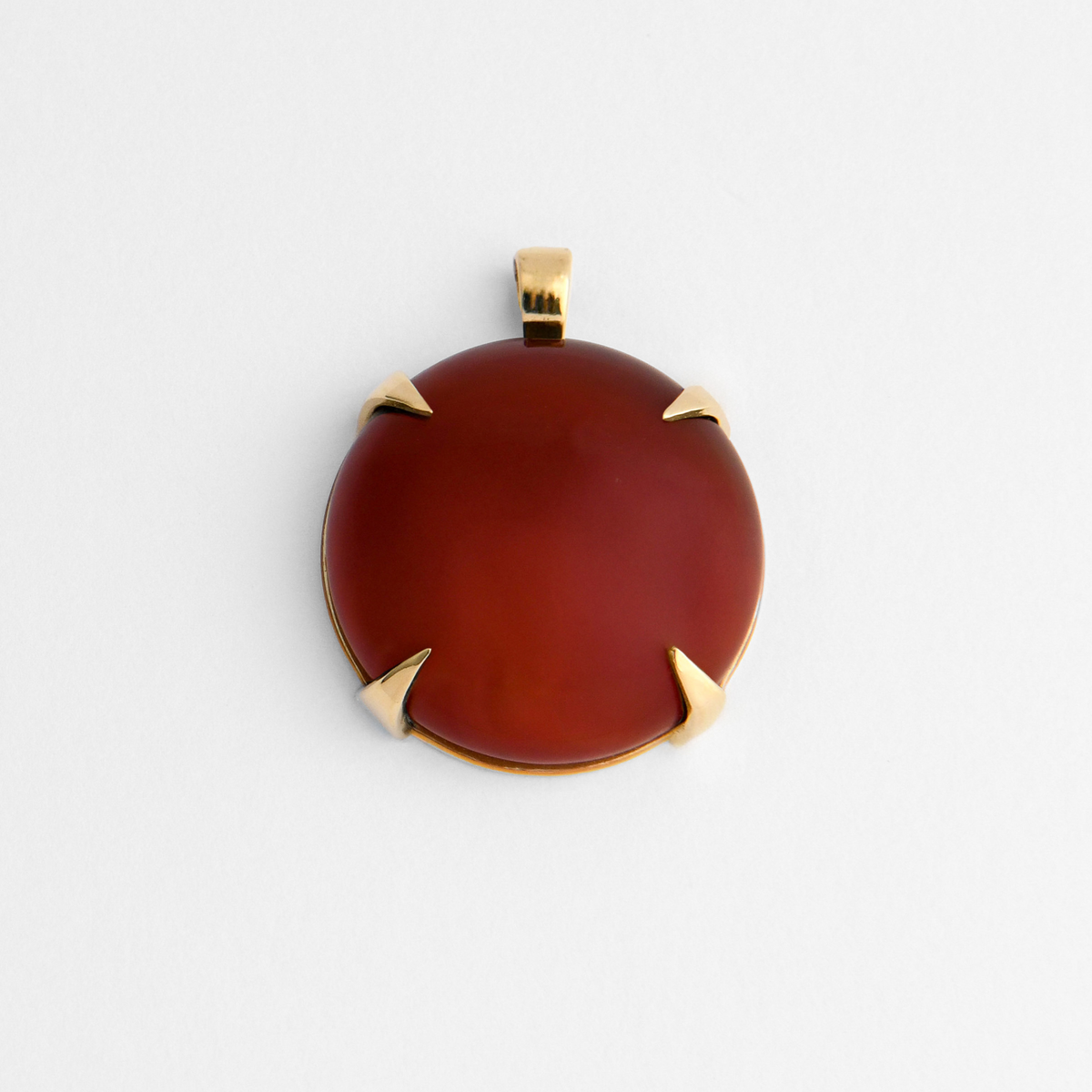 Spar, a round pendant with a small, squared bail, in warm, golden-yellow brass, bezel-set with deep reddish-brown red agate, ranging from dark burgundy to warmer amber-red where the light passes through. The natural stone varies in colour and markings from piece to piece.