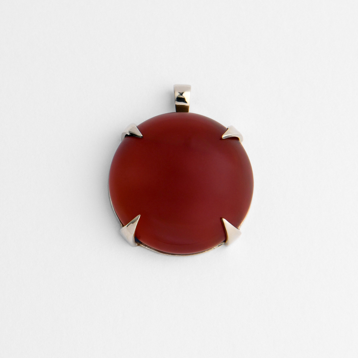 Spar, a round pendant with a small, squared bail, in cool, silvery white bronze, bezel-set with deep reddish-brown red agate, ranging from dark burgundy to warmer amber-red where the light passes through. The natural stone varies in colour and markings from piece to piece.