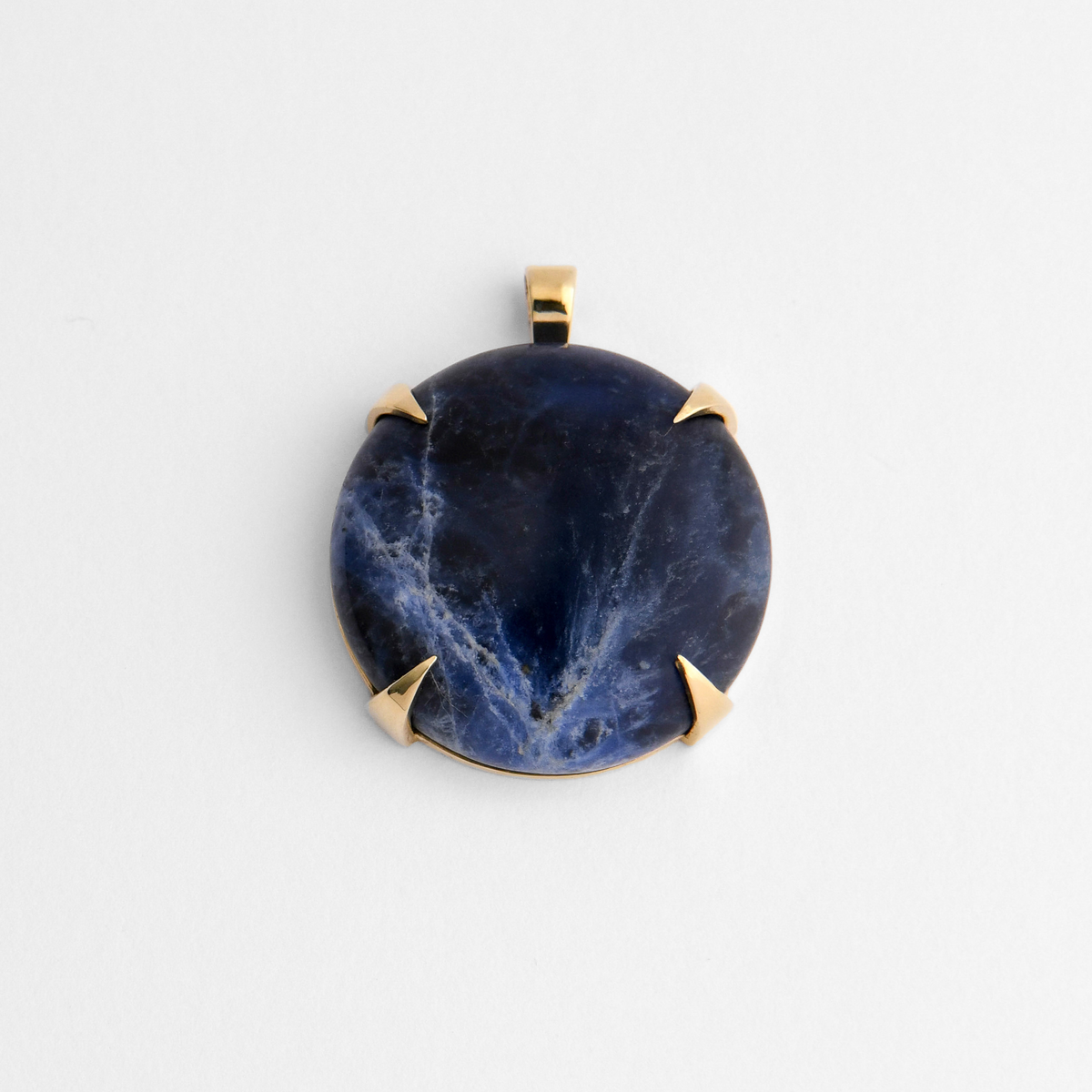 Spar, a round pendant in warm, golden-yellow brass, with four triangular claws holding blue sodalite, ranging from deep midnight to brighter blue, with irregular white veining and occasional biscuit-coloured markings. As a natural stone, each piece varies.