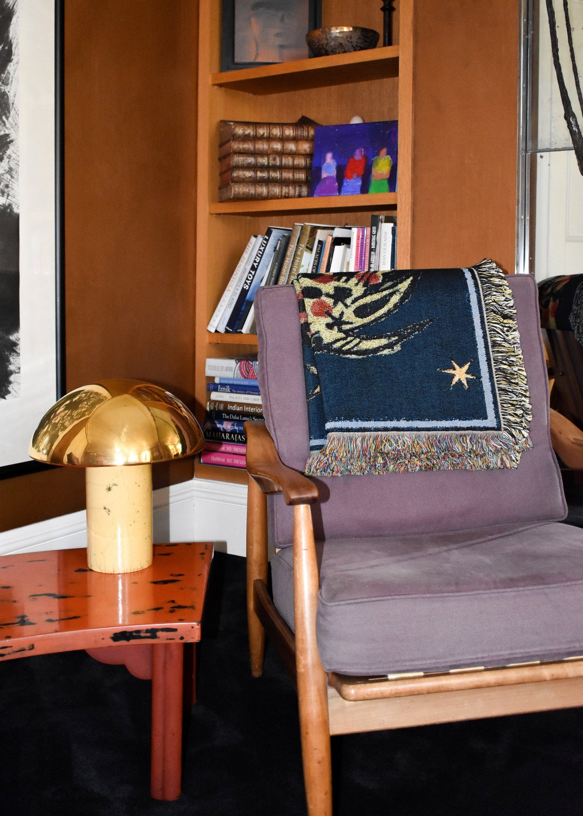 House of Hades Liminal Creatures cotton jacquard throw, folded over the back of a vintage wooden armchair upholstered in soft, dusty mauve velvet. The deep midnight-blue weave is decorated with golden stars, winding botanical motifs and touches of rich red, finished with a delicate pale blue border and thick, multicoloured fringe. Beside the chair, a polished brass mushroom-shaped lamp rests on a weathered terracotta-red table, while shelves of old leather-bound books, colourful artwork and collected obje