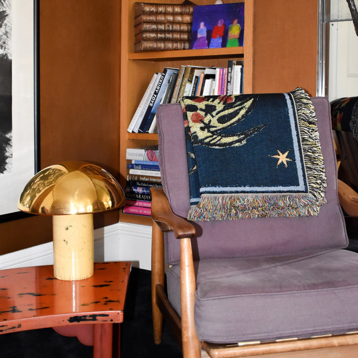 House of Hades Liminal Creatures cotton jacquard throw, folded over a vintage wooden armchair upholstered in dusty mauve. The midnight-blue weave features a golden star, winding botanical motifs in ochre and red, and a pale-blue border with multicoloured fringes. Beside the chair, a polished brass mushroom-shaped lamp rests on a weathered terracotta-red table, with shelves of leather-bound books and colourful artwork behind.