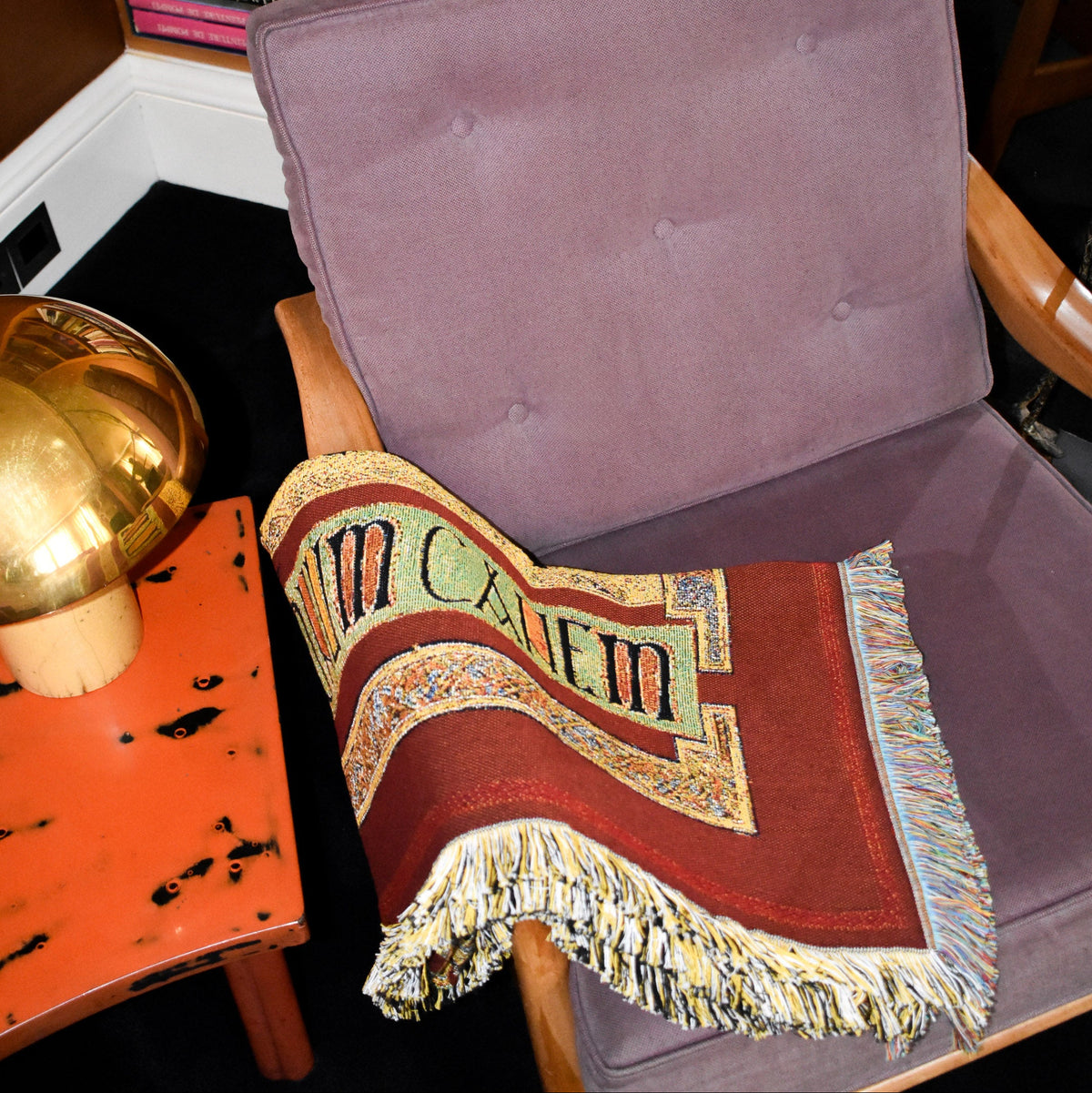House of Hades Beloved Beast cotton jacquard throw, loosely folded across a vintage wooden armchair upholstered in dusty mauve velvet. The rich oxblood-red weave features ornate golden borders and a pale sage-green panel with bold black Latin lettering, finished with thick cream, gold and multicoloured fringes. Beside the chair, a gleaming brass mushroom-shaped lamp rests on a weathered coral-red table, adding warmth to the richly textured interior.
