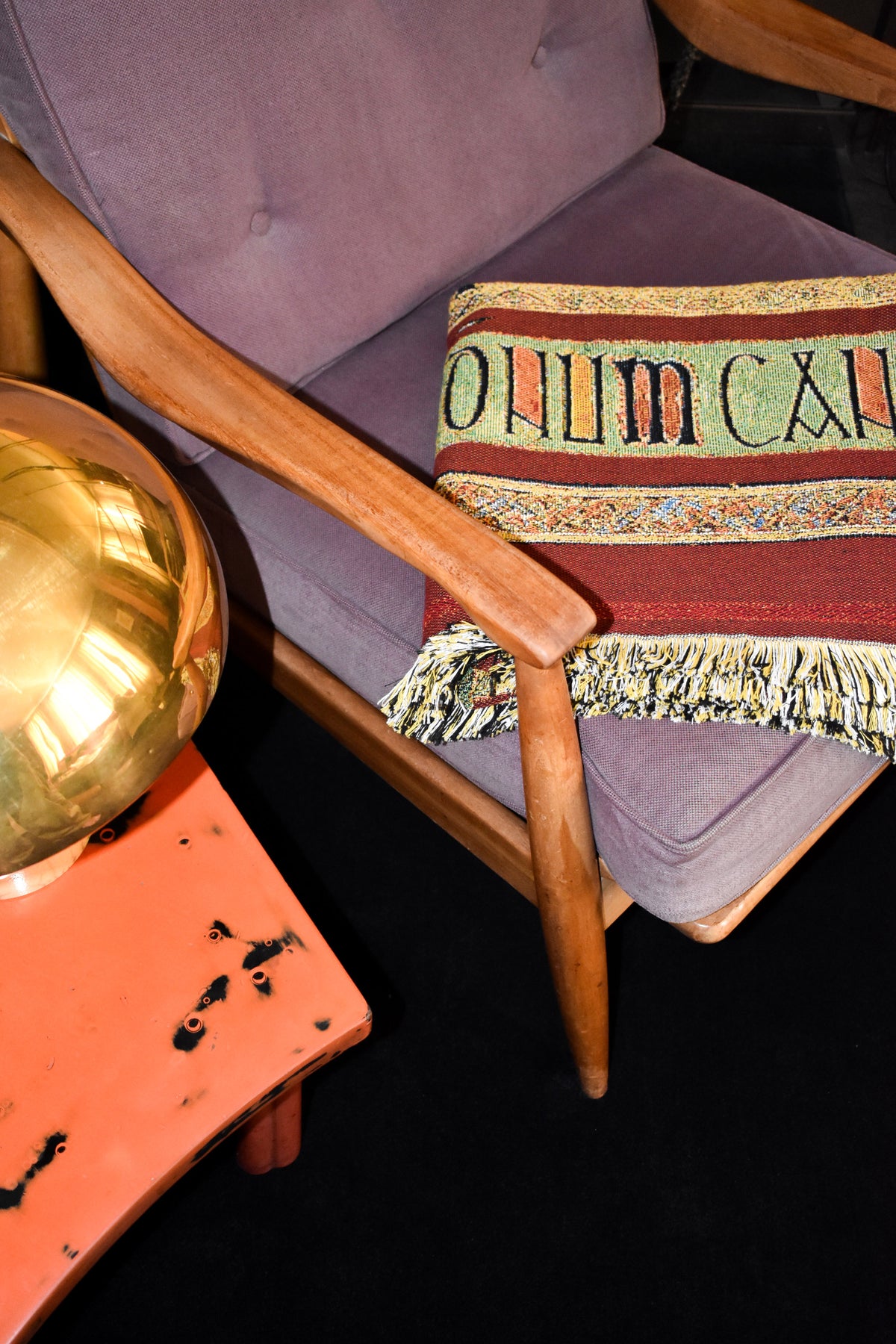 House of Hades Beloved Beast cotton jacquard throw, neatly folded across the seat of a vintage wooden armchair upholstered in dusty mauve. The rich oxblood-red weave reveals a decorative band of pale green, ochre and black lettering, framed by intricate patterned borders and a thick fringe of cream, black and golden threads. Beside the chair, a gleaming brass mushroom-shaped lamp sits on a weathered coral-red table, creating a warm, richly coloured interior.