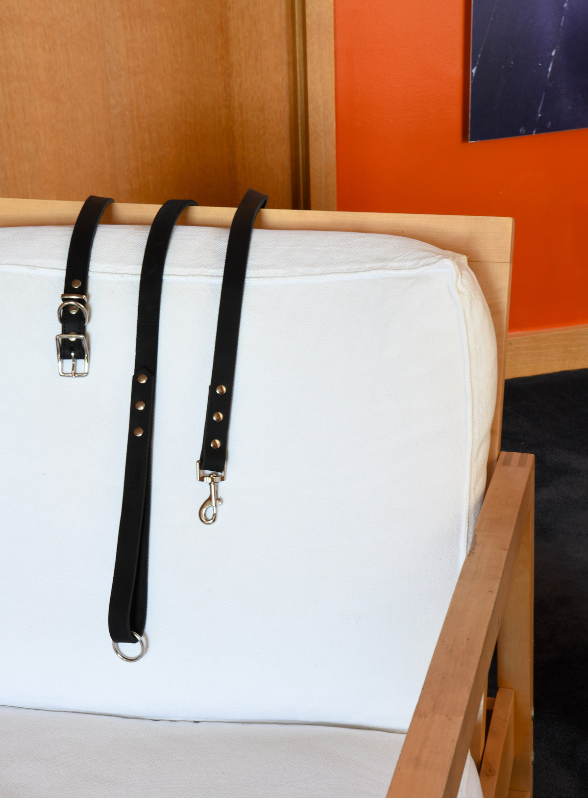 House of Hades black leather dog collar and matching lead with silver-coloured hardware, draped over the white cushioned back of a wooden armchair, against a vivid orange wall.