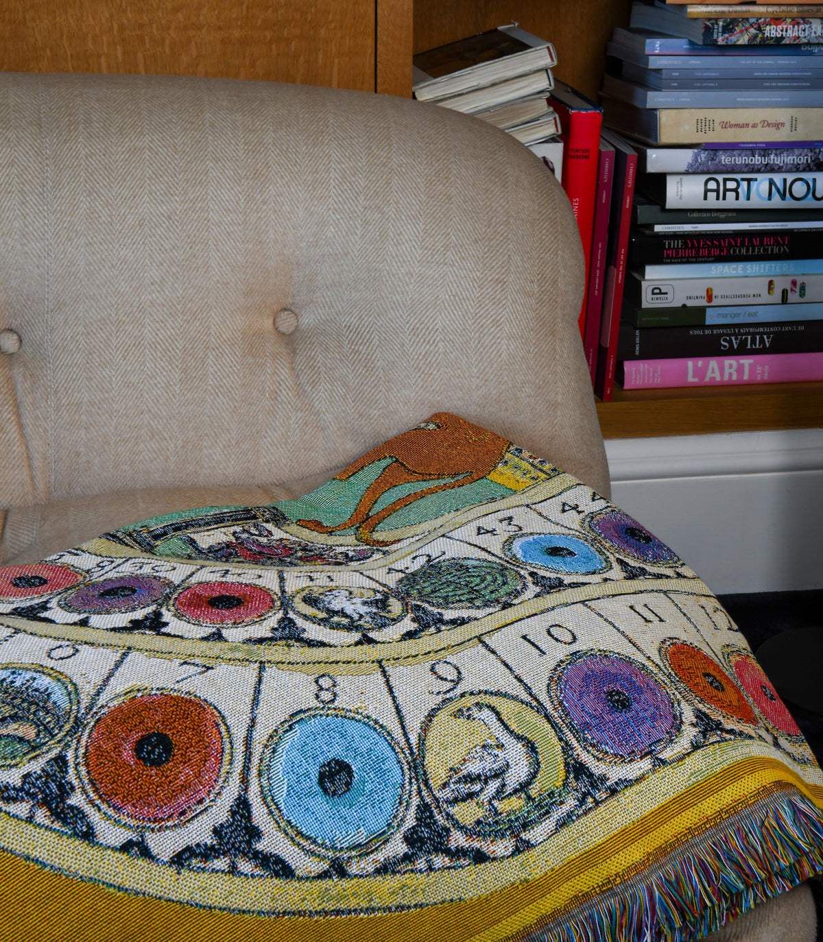 House of Hades Wayward & Wandering cotton jacquard throw, draped across a vintage cream herringbone armchair beside shelves of art books. The intricate woven design resembles an ancient celestial chart, with colourful circular motifs in turquoise, violet, orange and pink, mysterious illustrations and scattered numbers. A golden-yellow border and rich multicoloured fringe frame the tapestry-like fabric.