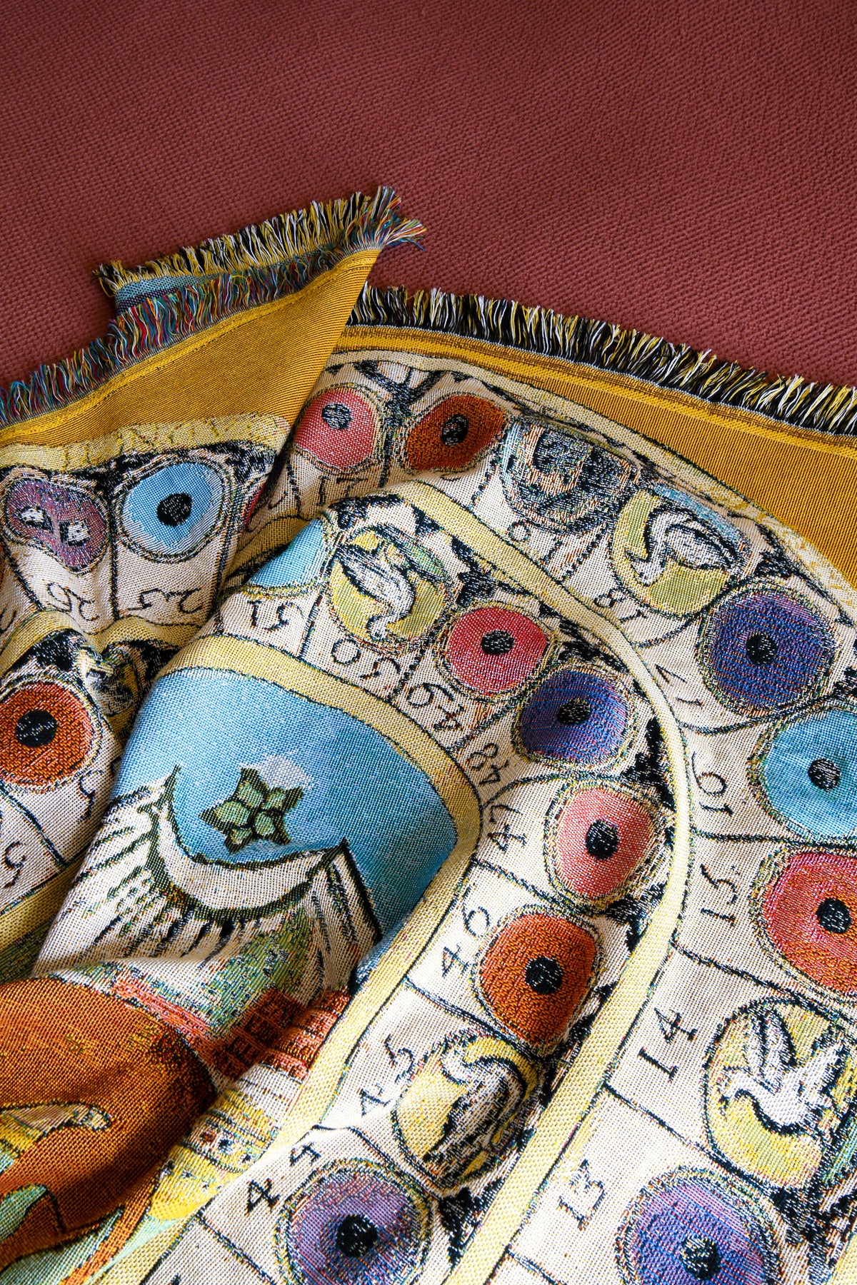 Close-up of the House of Hades Wayward & Wandering cotton jacquard throw, loosely folded against a rust-red background. The intricate woven design features colourful circular motifs, illustrated creatures and symbols in yellow, blue, orange and pink, with multicoloured fringed edges.