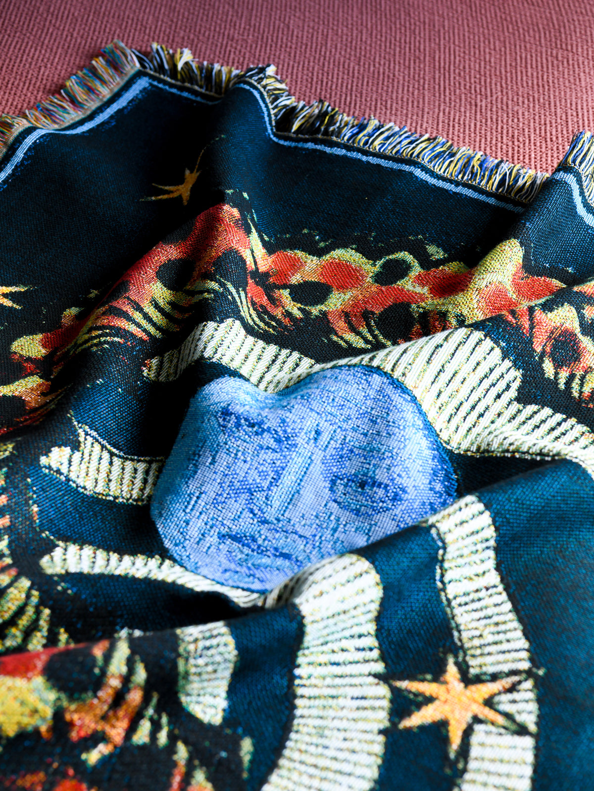 Close-up of the House of Hades Liminal Creatures cotton jacquard throw, gathered in soft, sculptural folds against a textured dusty-rose background. The deep midnight-blue weave reveals a striking cornflower-blue face with delicate, mysterious features, surrounded by winding ivory serpents, fiery red and orange botanical details, and scattered golden stars. A fine pale-blue border and richly coloured fringes frame the intricate design, revealing the texture and detail of the woven threads.
