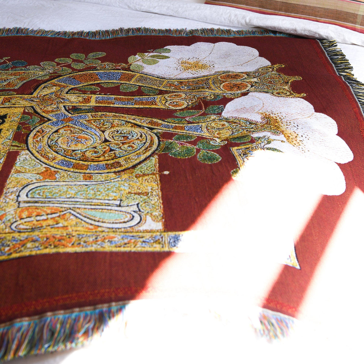 Close-up of the House of Hades Beloved Beast cotton jacquard throw laid across a white bed, its rich oxblood-red background adorned with large ivory flowers, delicate green leaves and elaborate golden scrollwork with touches of blue and ochre. Intricate ornamental lettering and multicoloured fringed edges frame the tapestry-like design, while a bright shaft of sunlight falls diagonally across the fabric, illuminating its woven texture.