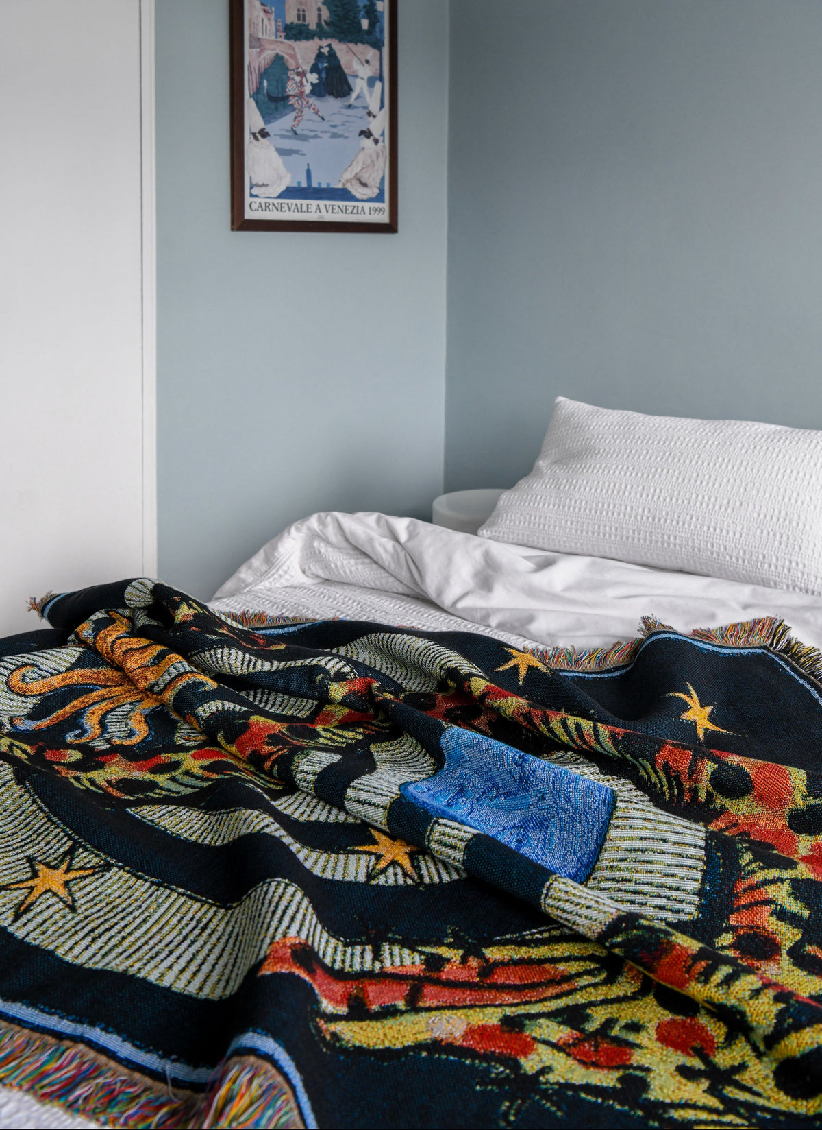 House of Hades Liminal Creatures cotton jacquard throw, loosely draped across a white bed in a pale blue bedroom. The dark navy woven design features mythical creatures, winding serpents and golden stars, with colourful detailing and fringed edges. In the background is a framed poster of the Carnival of Venice set at night sharing similar colours.