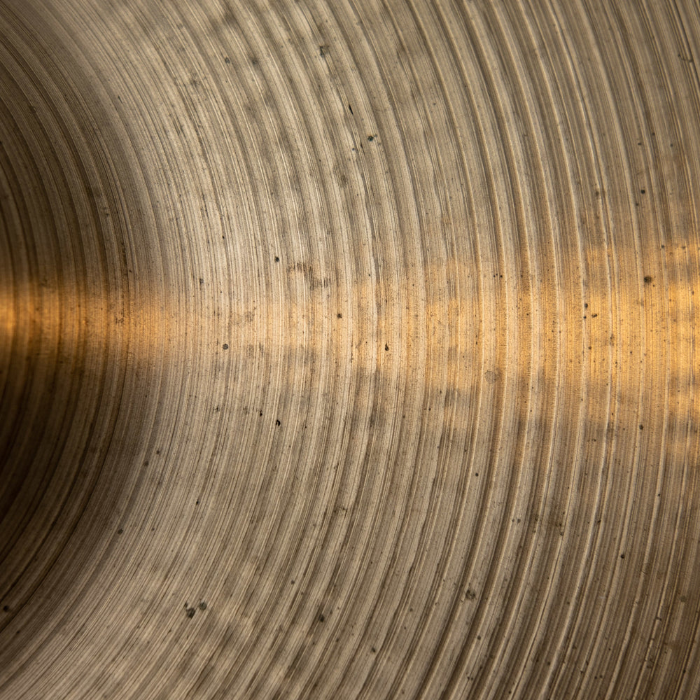 A cropped close-up of an aged brass cymbal. Concentric ridges create waves of light and shadow, with a ghost of golden light smoking through the centre.